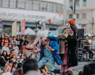 En una plaza llena de público, un payaso carga en brazos a otra artista mientras una tercera, de abrigo de leopardo, mira la escena.