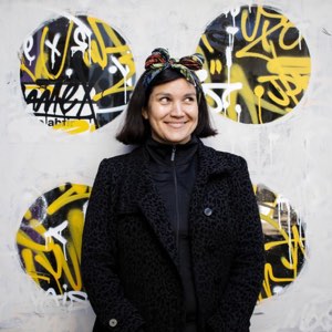 Katherine Cisternas apoyada en el mismo muro pintado, con una pañoleta anudada en el pelo y un abrigo negro.