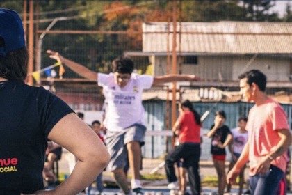 Un niño camina con los brazos abiertos sobre una barra de equilibrio, en una cancha de barrio, mientras una monitora con polera de Quetralmahue lo observa de espaldas.