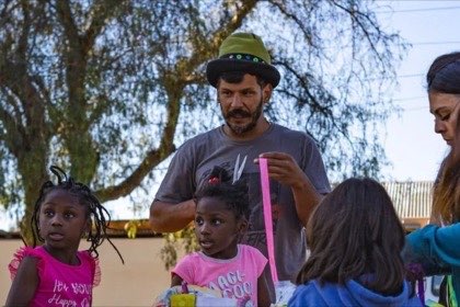 Cristian Segovia, de sombrero verde, explica algo a un grupo de niñas alrededor de una mesa, al aire libre.
