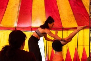 Sue Abarca sostiene las piernas de una participante que hace una parada de cabeza, dentro de una carpa a rayas rojas y amarillas.