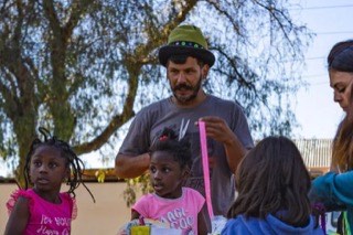 Cristian Segovia, de sombrero verde, explica algo a un grupo de niñas alrededor de una mesa, al aire libre.