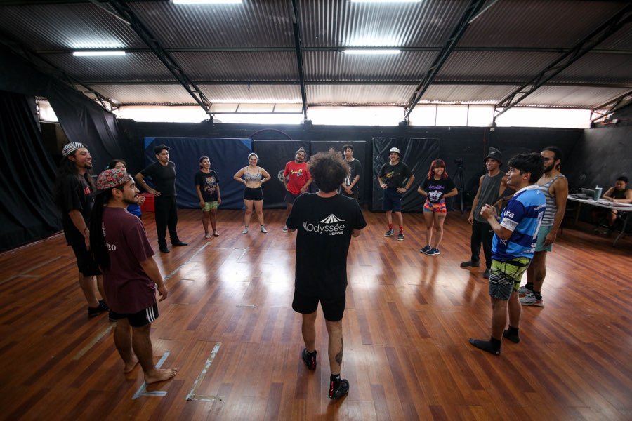Una rueda de participantes de pie en una sala de ensayo de piso de madera, escuchando al profesor que está en el centro, de espaldas.